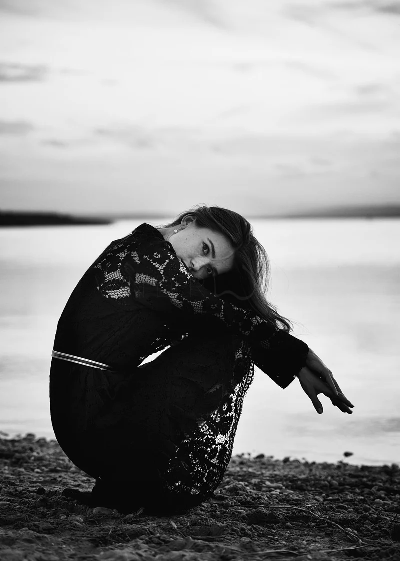 Black and white photo of a woman with long hair in a lacy dress, squatting by the water's edge with her arms resting on her knees, looking at the camera with a serious expression.