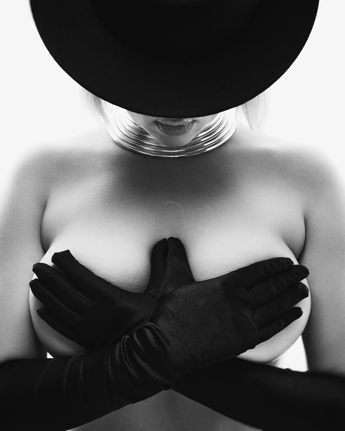 Black and white photo of a woman wearing a wide-brim hat, large statement necklace, and gloves, covering her chest with her hands.