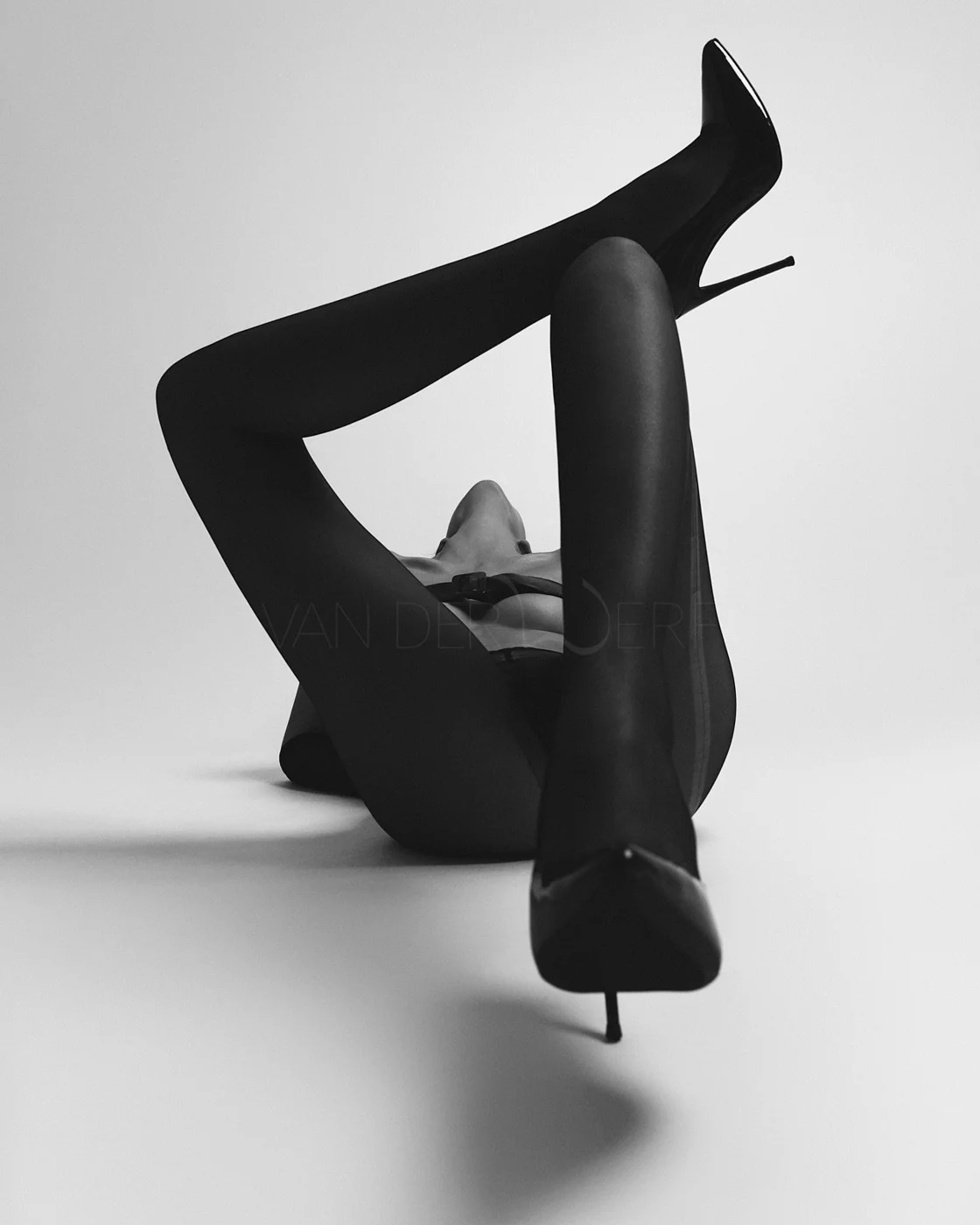 A woman lying on her back with one leg raised and bent, wearing high-heeled shoes and sheer tights, black and white photograph against a plain background.