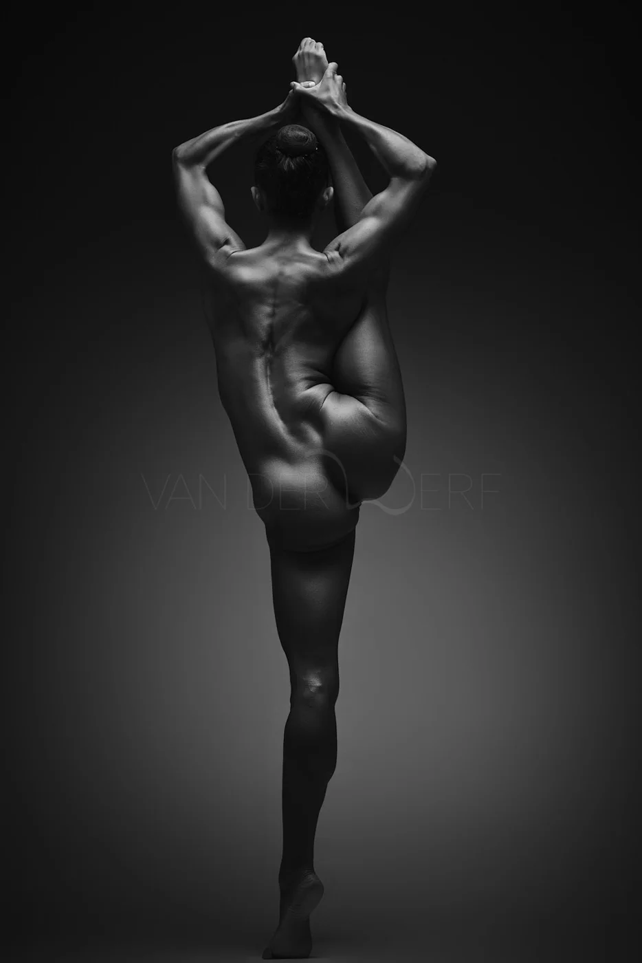 A black-and-white photo of a nude woman performing a yoga pose, balancing on one leg with arms raised and hands clasped above her head.