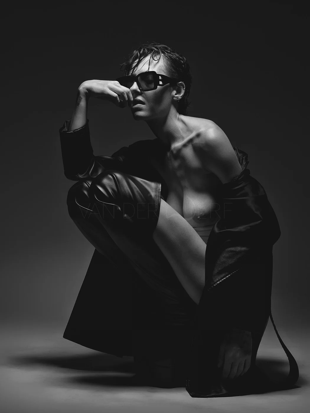 A black-and-white photograph of a person with short hair wearing sunglasses, a leather jacket, and revealing clothing, posing against a dark background.