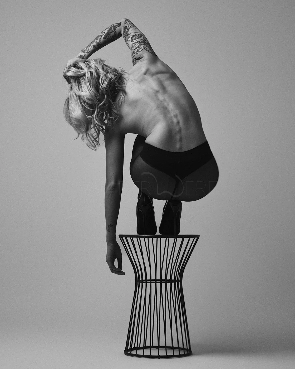 A woman with blonde hair and tattoos crouches on a wire stool, balancing on her toes, with her back arched and her left arm raised above her head, in a black and white studio shot.