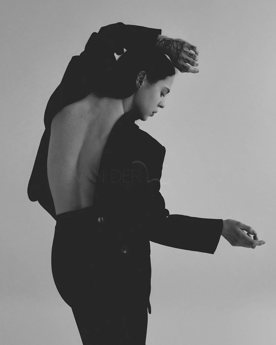 A black and white photo of a woman with short hair, partially dressed in a black blazer, with her back and shoulder exposed, and her right arm raised above her head. She appears to be looking down with a contemplative expression.
