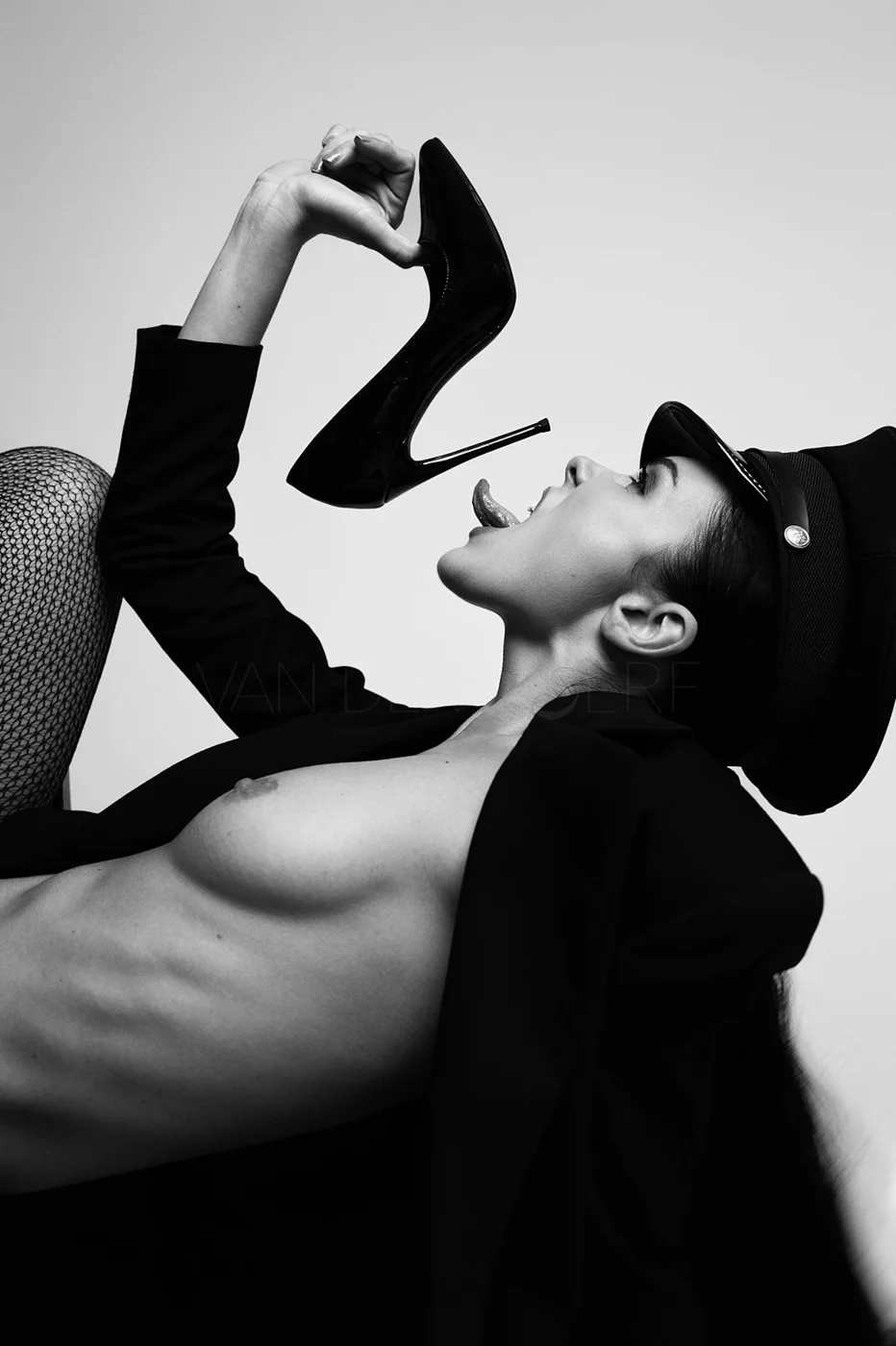A black-and-white photo of a woman lying down, wearing fishnet stockings, a black jacket, and a black hat. She is holding a high-heeled shoe near her face with her mouth open and tongue out.