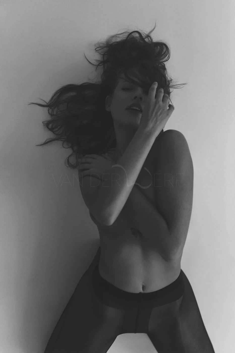 A black-and-white photo of a woman with long, wavy hair lying on a surface, partially covering her face with her hand, wearing sheer black leggings and a top that exposes her midriff.