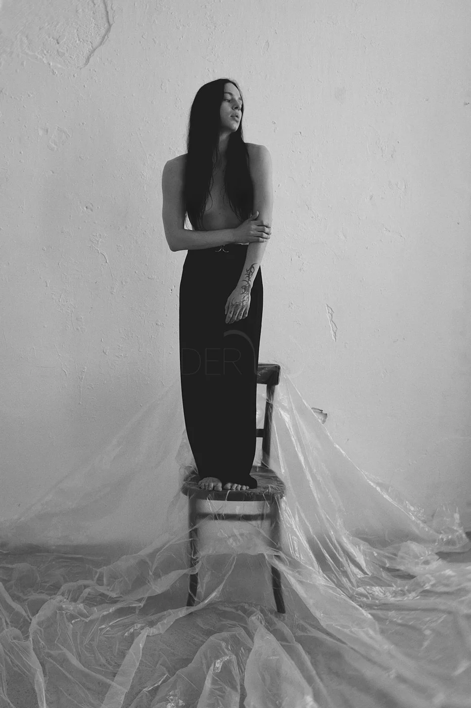 A black and white photo of a woman with long dark hair standing on a chair covered with plastic, against a plain wall. She is topless, wearing black pants, and appears to be posing thoughtfully with one arm crossed over her chest and hand on her oppo