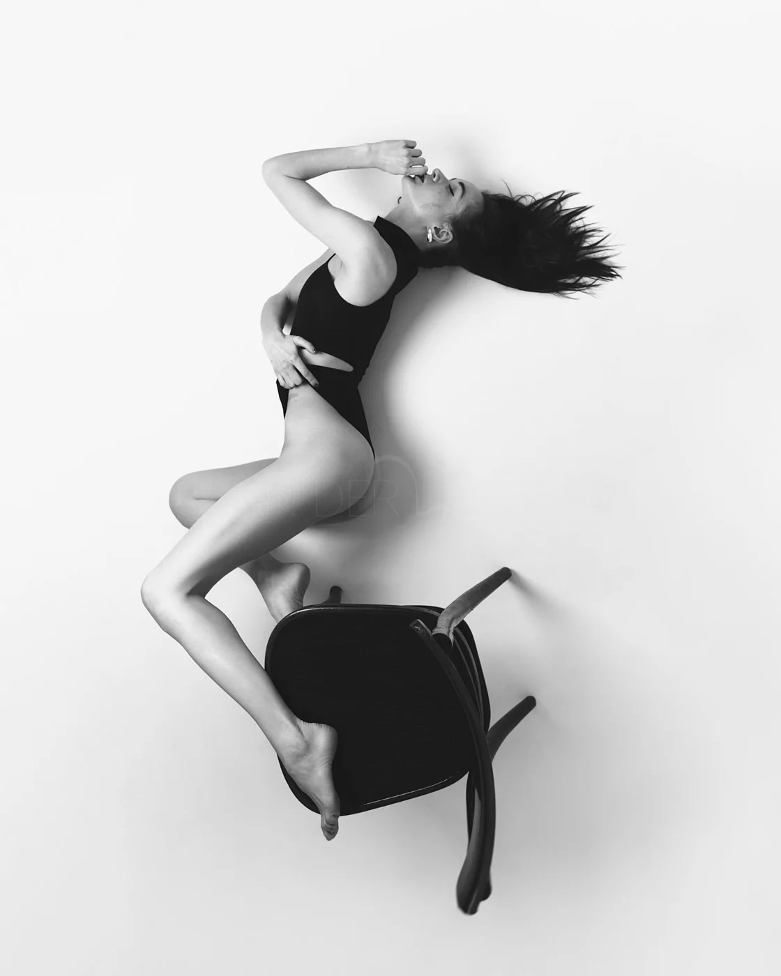 A woman in black lingerie lying on a chair with her head tilted back, hair spread out, and one hand on her stomach while the other touches her lips, against a plain white background.