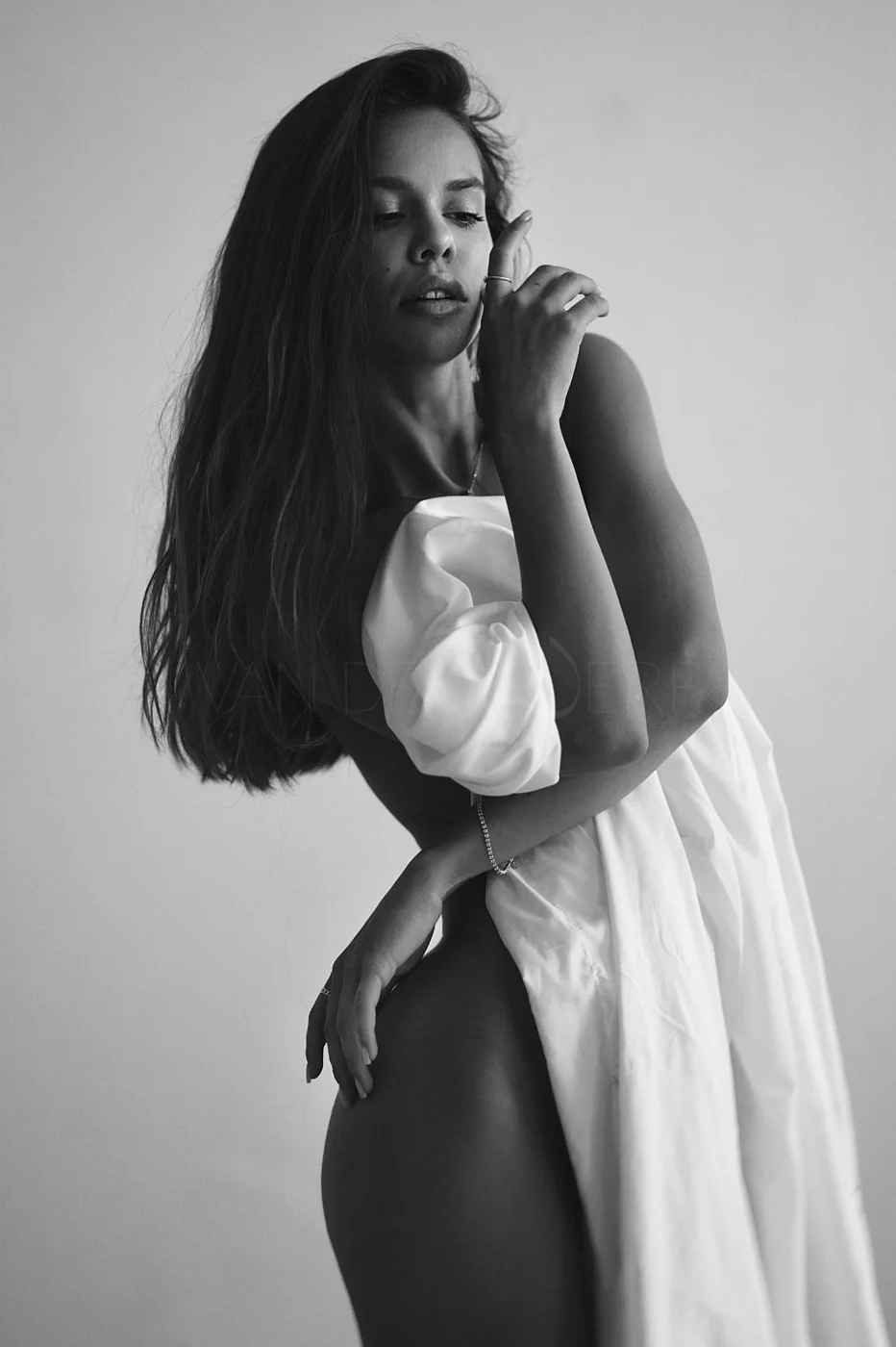 A black-and-white portrait of a woman with long wavy hair, wearing a white garment off her shoulder, with one hand on her chest and the other on her hip, looking contemplative.
