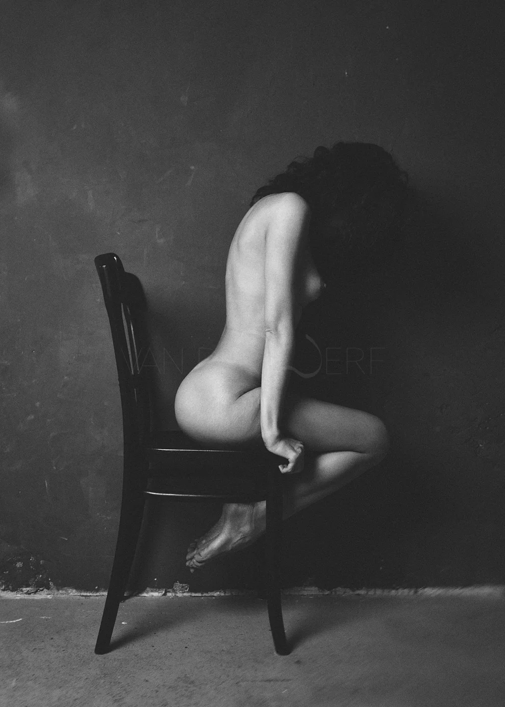 A black and white photograph of a nude woman sitting sideways on a wooden chair against a dark background. Her head is bowed, and her hair covers her face.