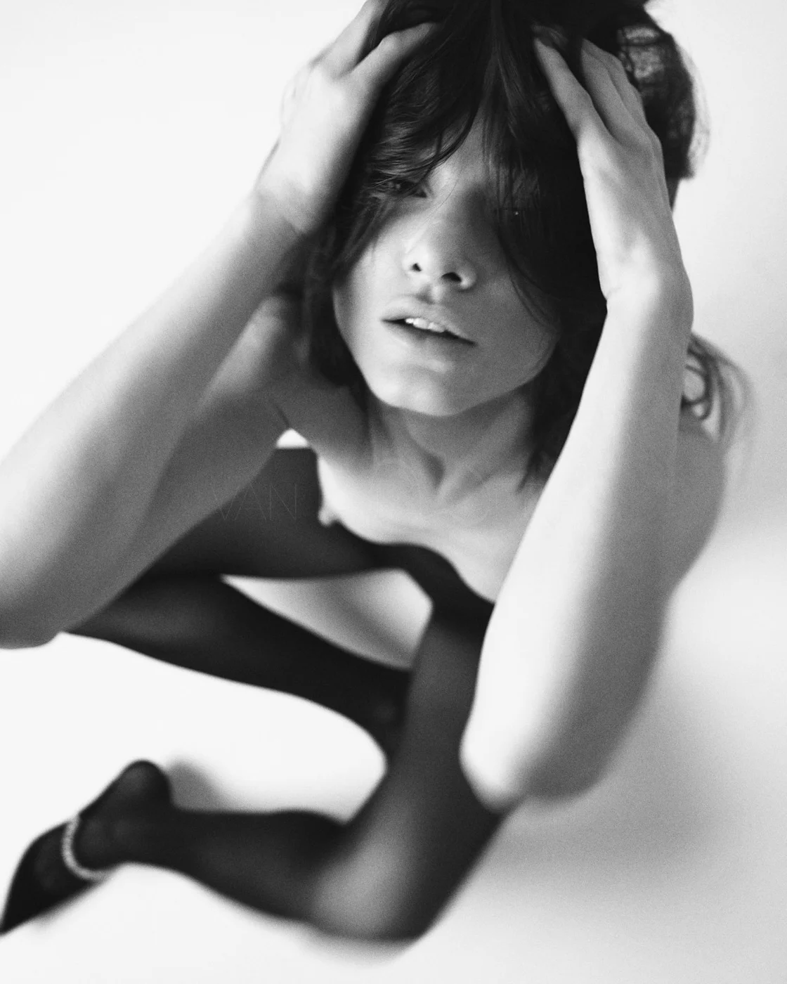 A black and white photo of a woman with dark hair, holding her head with both hands, looking slightly upward with a relaxed expression.
