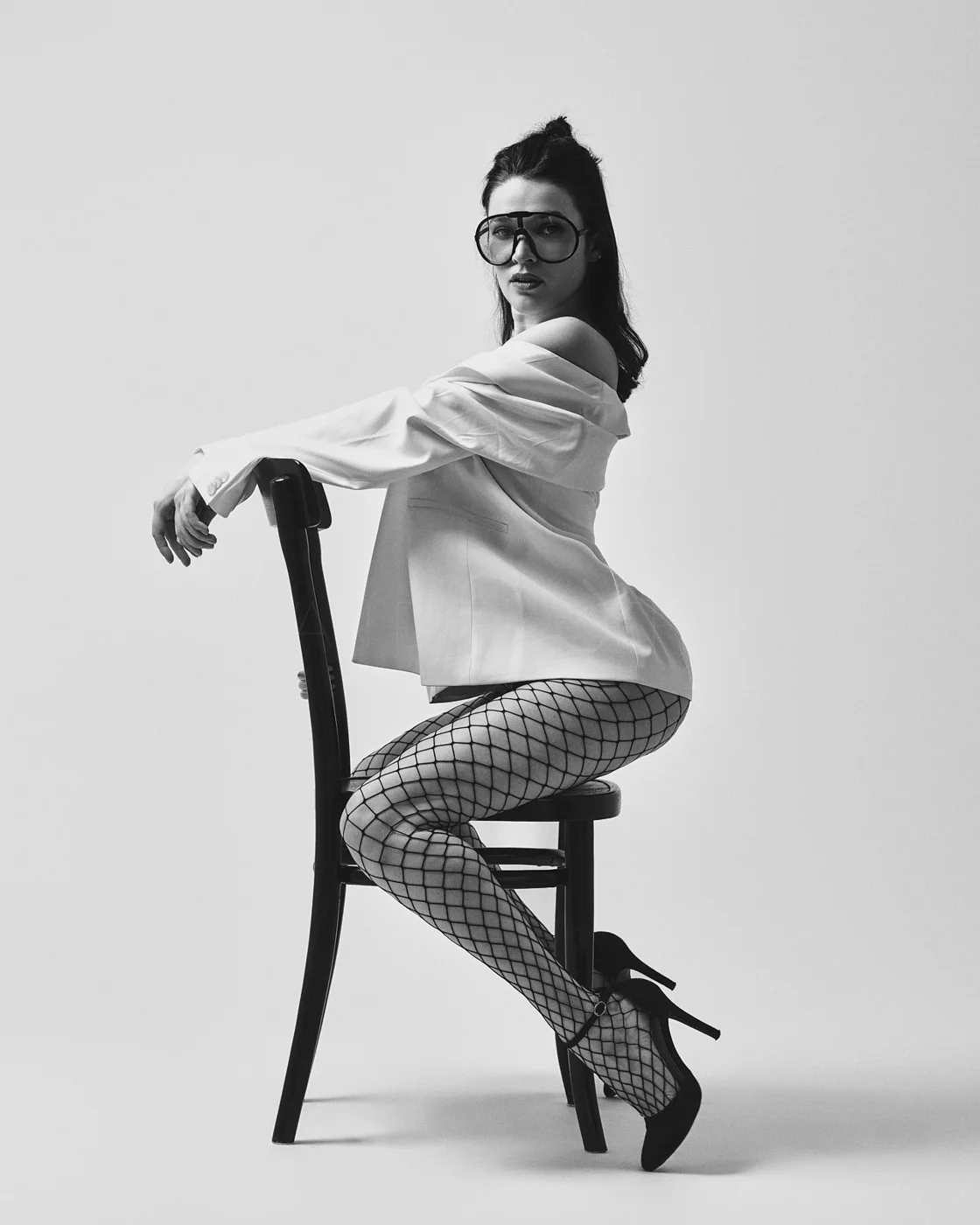 Black and white photo of a woman sitting sideways on a chair, wearing glasses, an off-the-shoulder blazer, fishnet stockings, and high heels, with a plain background.