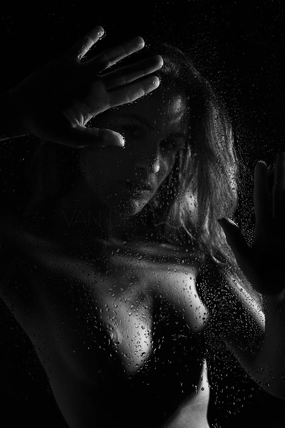 A black and white photo of a woman behind a rainy glass, with her hand touching the glass and water droplets on the surface.