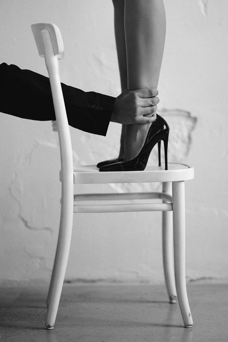 A person is seated on a white wooden chair wearing black high heels, while another person in a suit is holding their ankle.