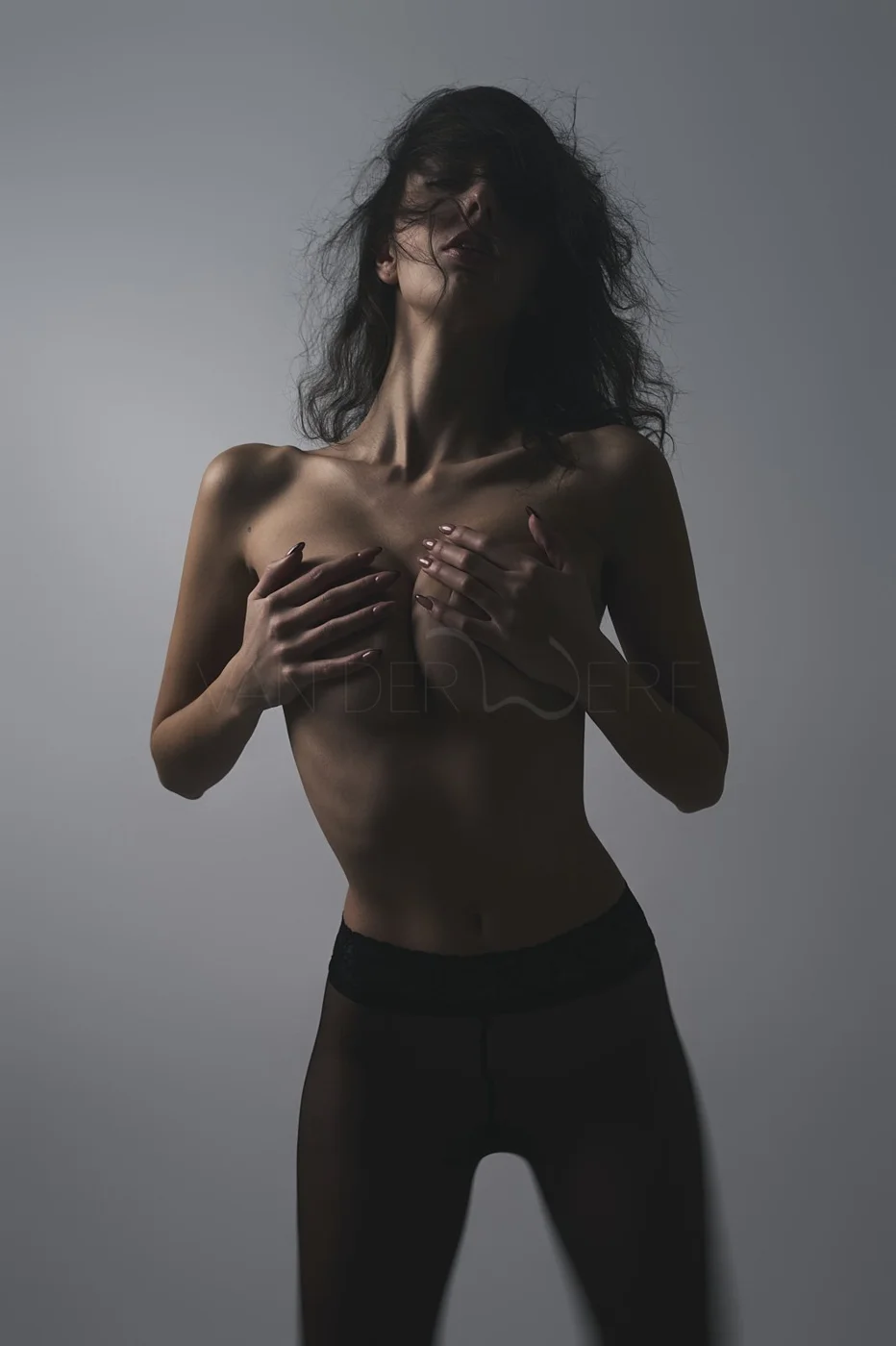 A woman with dark, tousled hair poses topless, covering her chest with her hands, wearing black lace stockings against a gray background.