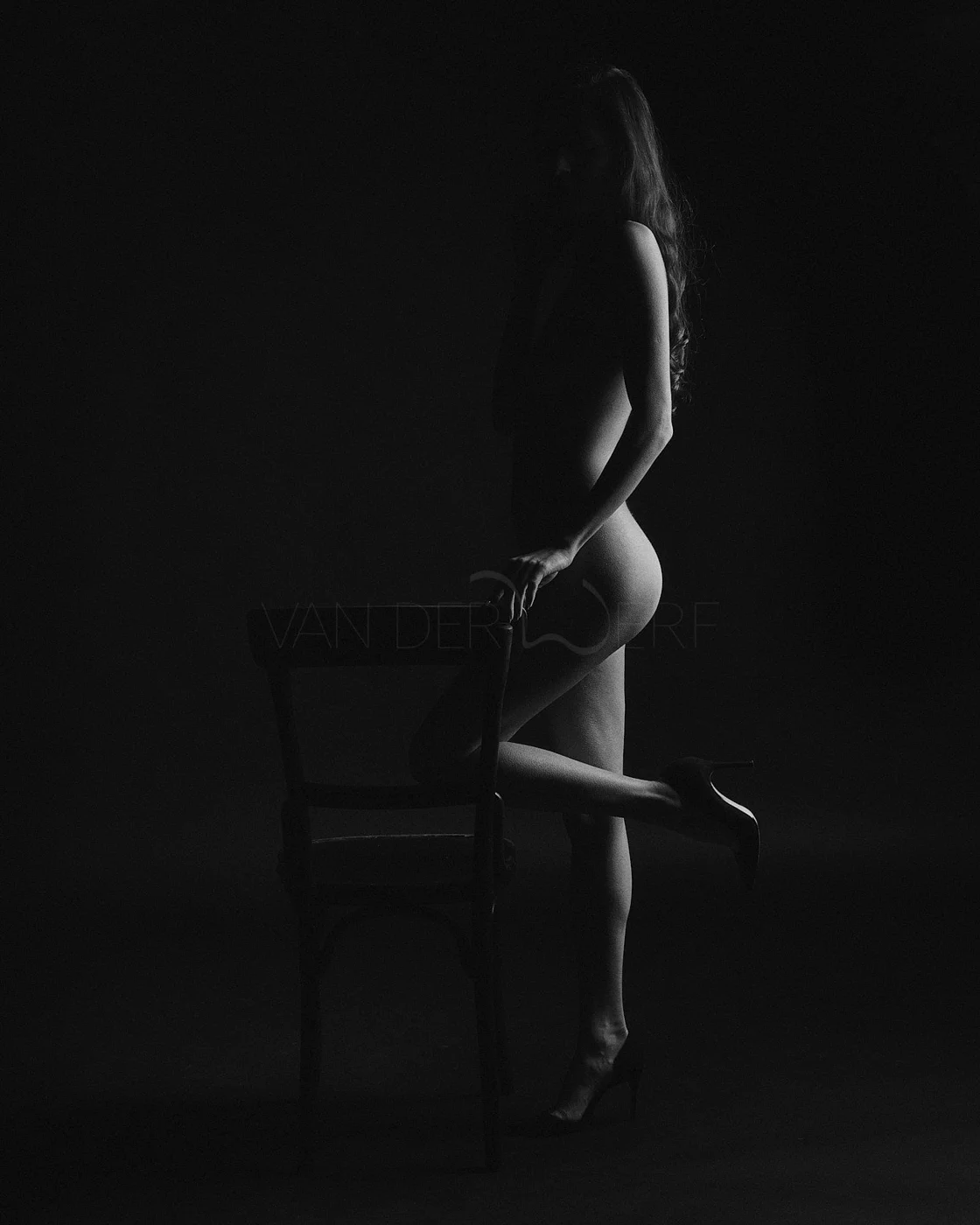 A black and white silhouette of a woman standing behind an empty chair, with one leg raised and her hand resting on the chair, in a dark studio setting.