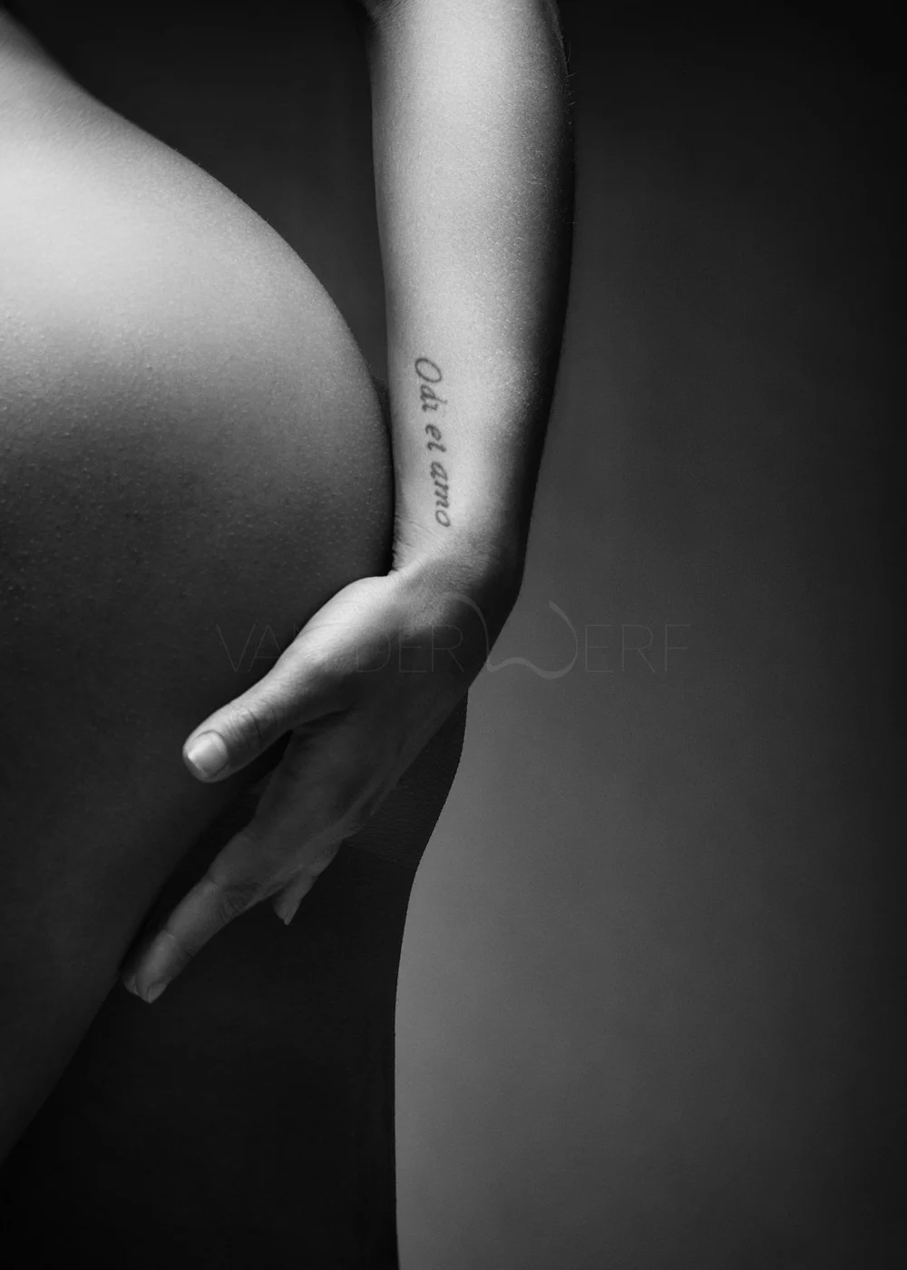 Close-up black and white photo of a person's hand resting on their lower back, with a tattoo that reads 'Dare to' on the inner forearm.