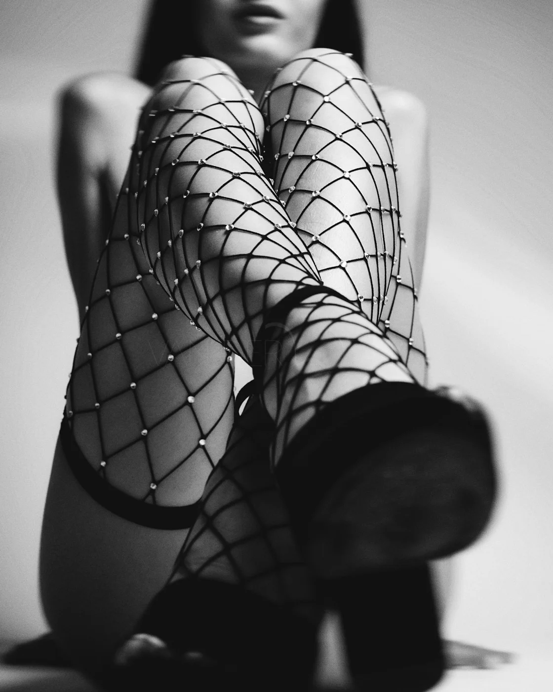 Black and white photo of a woman wearing fishnet stockings and high heels, sitting with her legs crossed and the camera focuses on her legs.