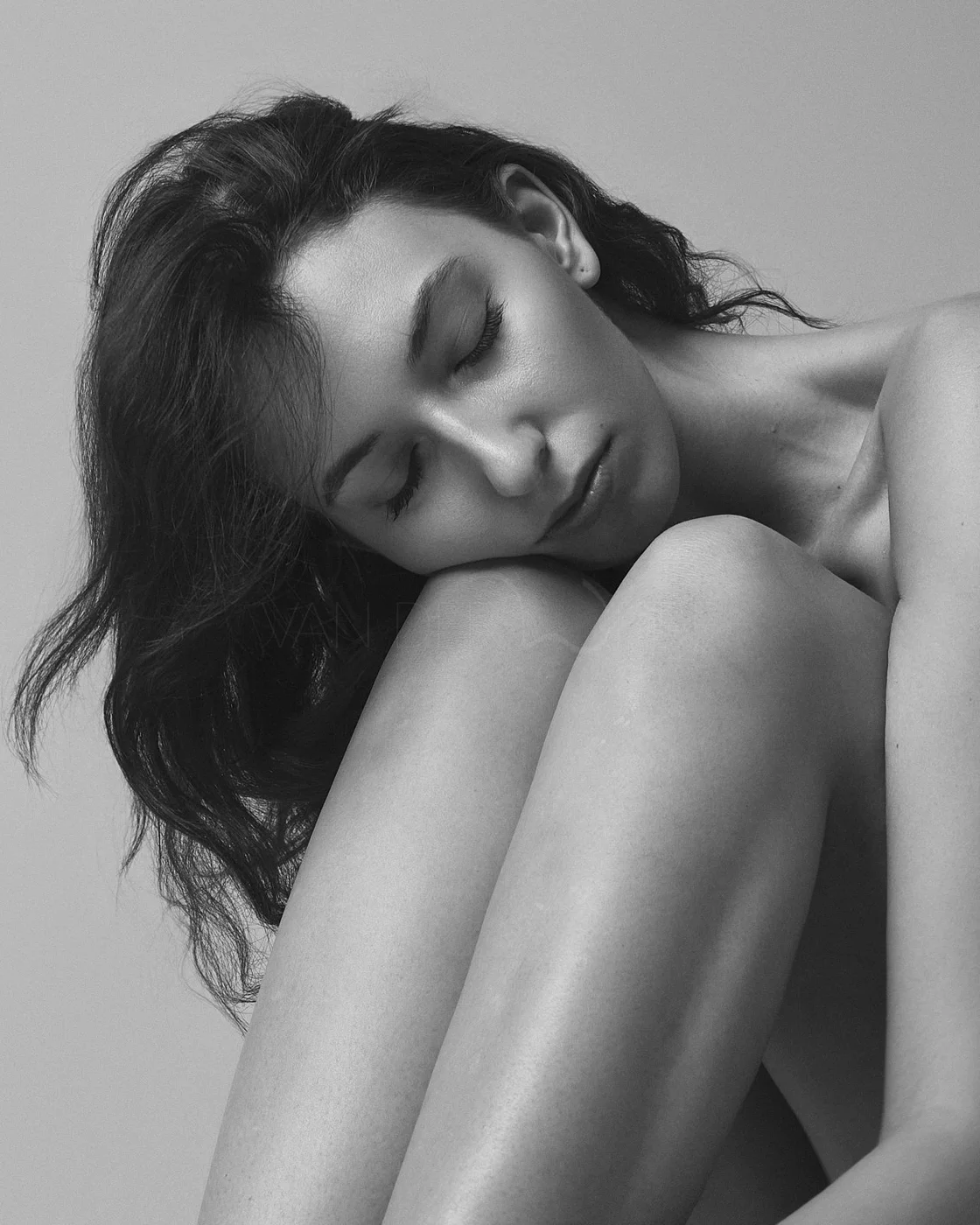 Black and white portrait of a woman with closed eyes, resting her head on her knee, with dark wavy hair and smooth skin.