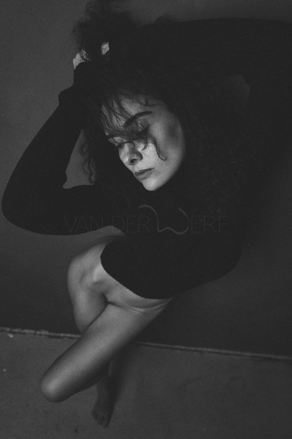 Black and white photo of a woman with curly hair, wearing a black long sleeve shirt, posing with her eyes closed and her hand touching her head, against a dark background.