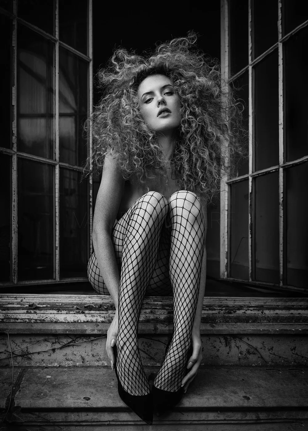 A black and white photo of a woman with curly hair, sitting on a window sill, wearing fishnet stockings and high heels, looking directly at the camera with a serious expression.