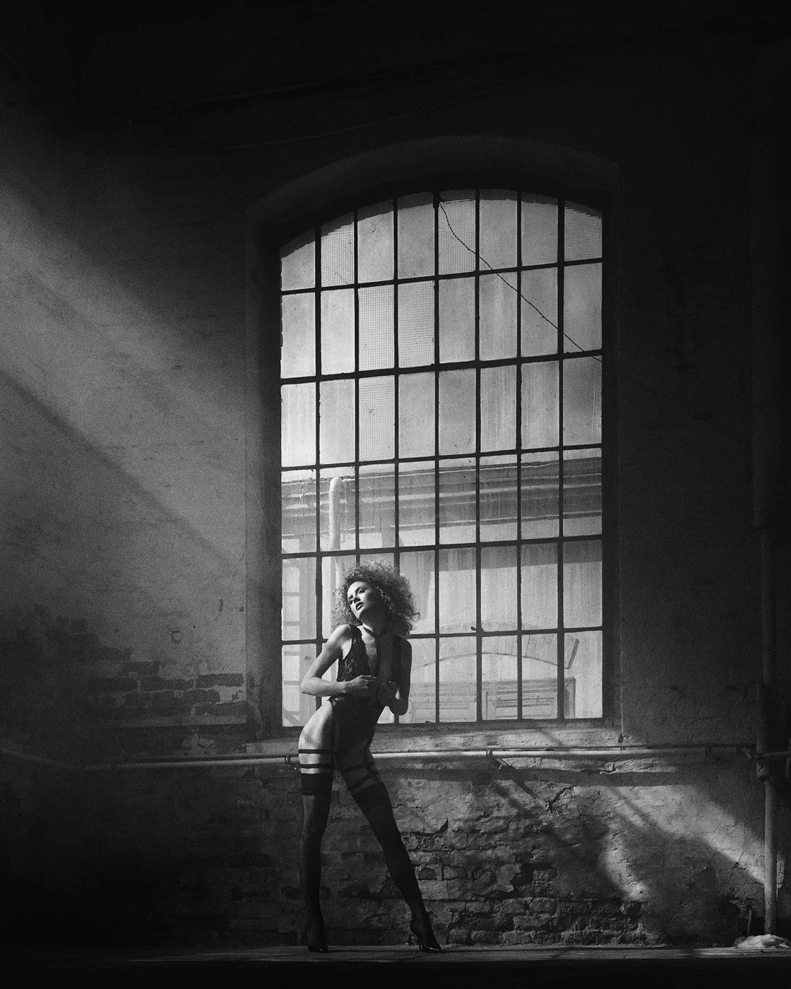 A woman with curly hair in black lingerie and thigh-high stockings stands in front of a large industrial window in a dark, weathered room, with dramatic light and shadows.