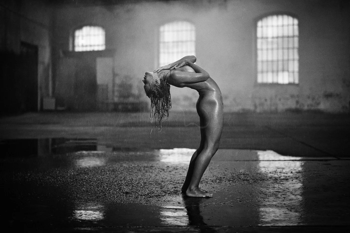 A woman standing wet in an industrial-style room with high windows, arch-shaped panes, covering her face with her hands and arching her back.