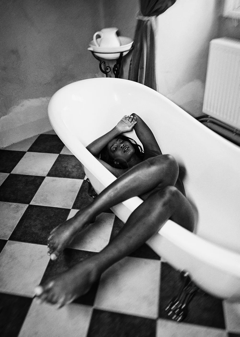A woman is lying in a vintage-style bathtub with her eyes closed, resting her head on her arm. The bathroom has black and white checkered flooring with a side table holding a teapot and cups, and the scene is captured in black and white.