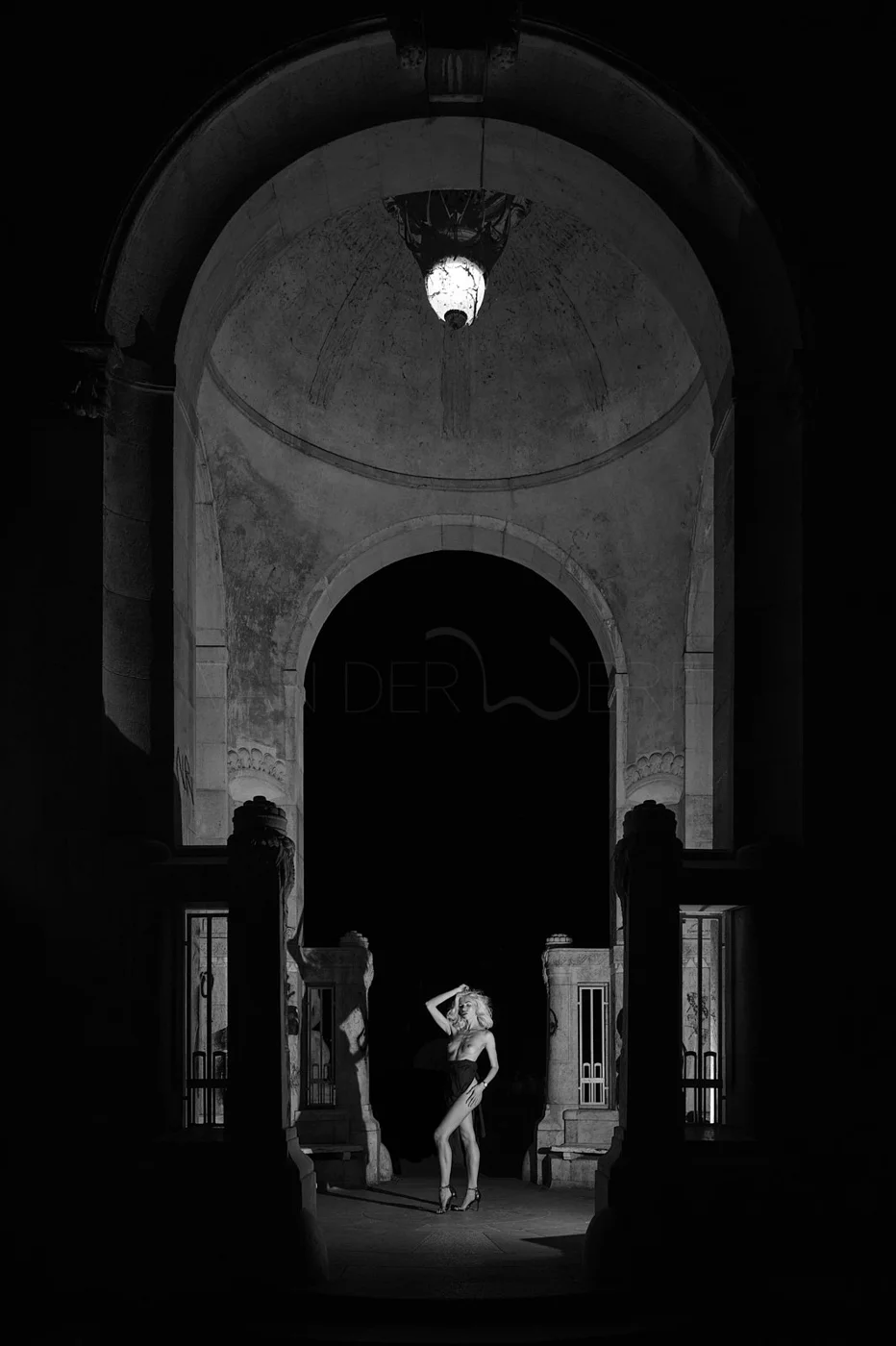 A black and white photo of a woman with blonde hair standing in an arched, ornate architectural setting, illuminated by a hanging light fixture, with shadows cast around her.