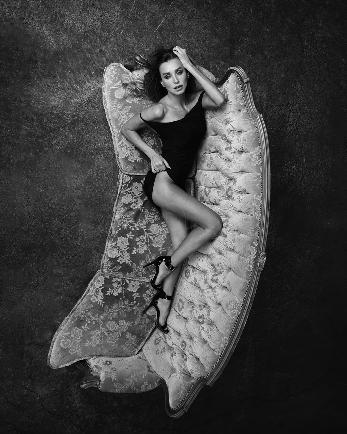 A woman with long hair lying on a vintage floral upholstered sofa, wearing a black dress and high heels, in black and white photography.
