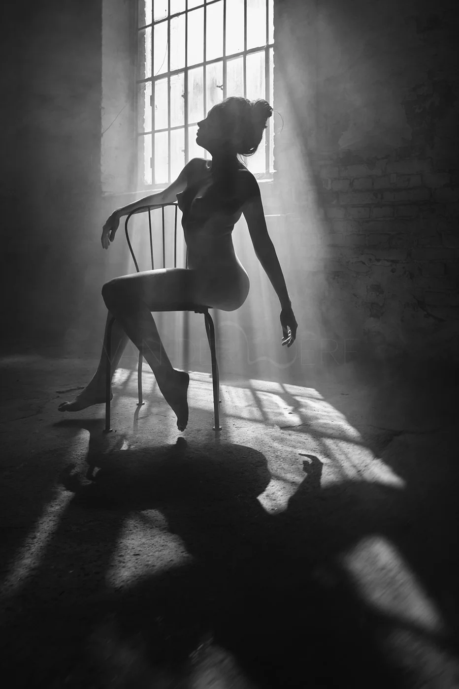 Silhouette of a nude woman sitting on a chair in a room with brick walls and a large window, with sunlight streaming in creating dramatic shadows.