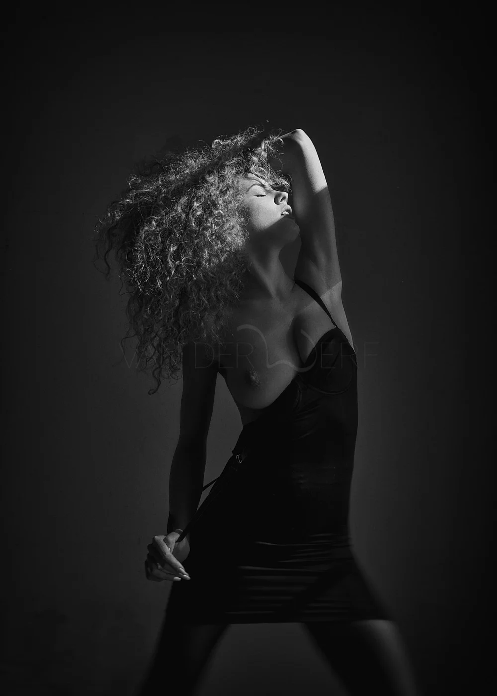 Black and white photo of a woman with curly hair posing with one arm over her head and eyes closed, wearing a black dress with one strap. The background is dark.