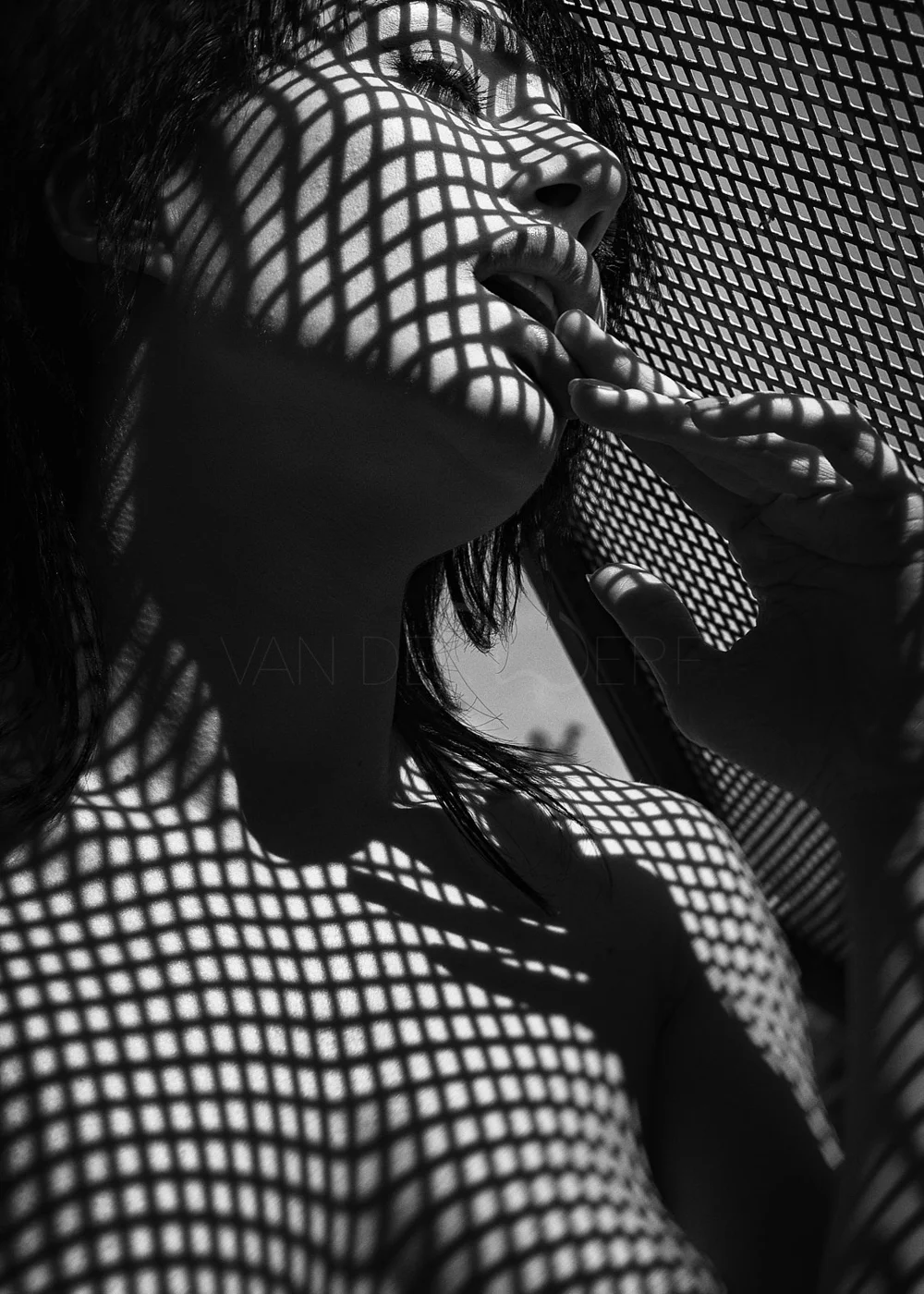A black and white photo of a woman with shadows of a patterned surface cast across her face and body, creating a grid-like design.