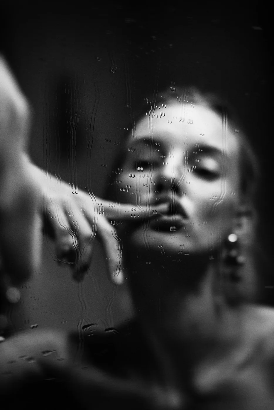 A black-and-white photograph of a woman looking at her reflection through a rain-covered window. Her face appears blurred and mysterious.