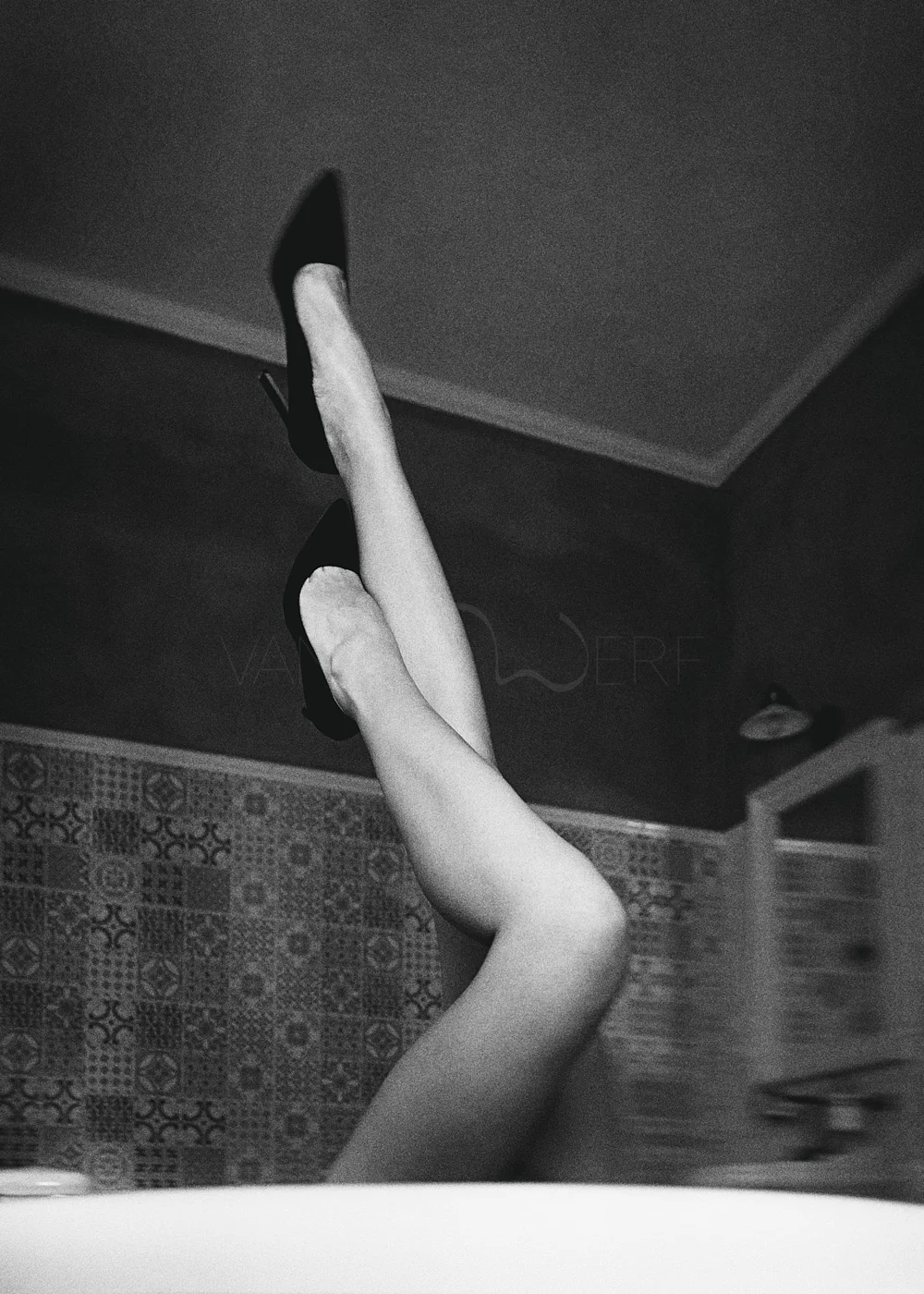 A black-and-white image showing a person's legs raised in the air, wearing high heel shoes, in a room with patterned wallpaper and a ceiling light.