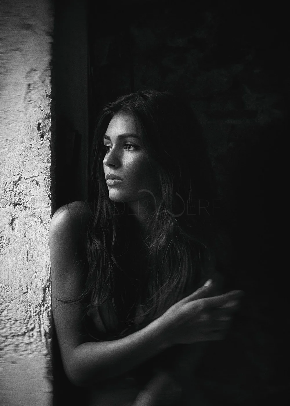 A black and white portrait of a woman with long hair, gazing thoughtfully out of frame, sitting by a textured wall, with low lighting on her face and arm.