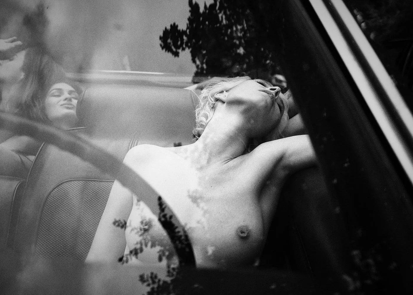 Black and white photo of a topless woman with light-colored hair, lying on her side in the back seat of a car, with another woman visible through the window, looking at the camera.