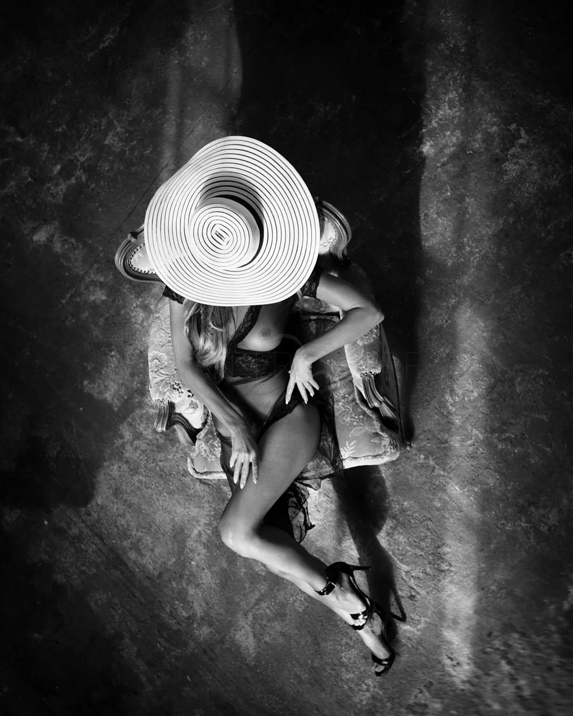 Black and white overhead view of a woman reclining on an ornate chair, wearing a wide-brimmed hat, lingerie, and high heels.