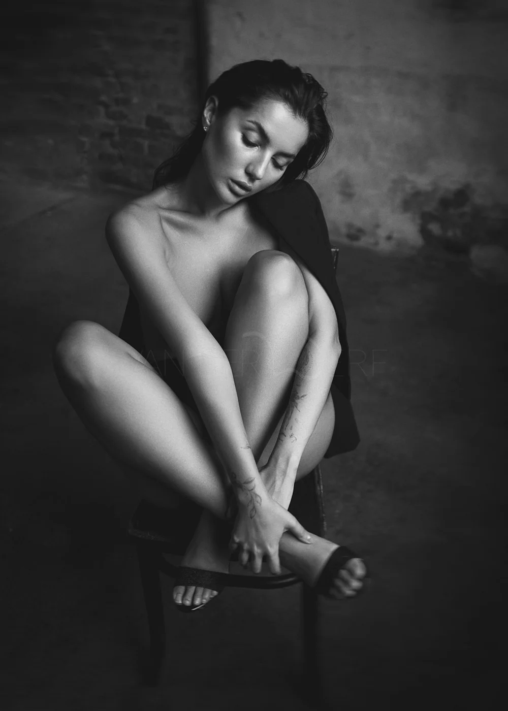 Black and white photo of a woman sitting on a chair, with her knees bent and arms wrapped around her legs. She has long dark hair, tattoos on her arms, and is wearing sandals and a blazer draped over her shoulders, looking down with a contemplative e