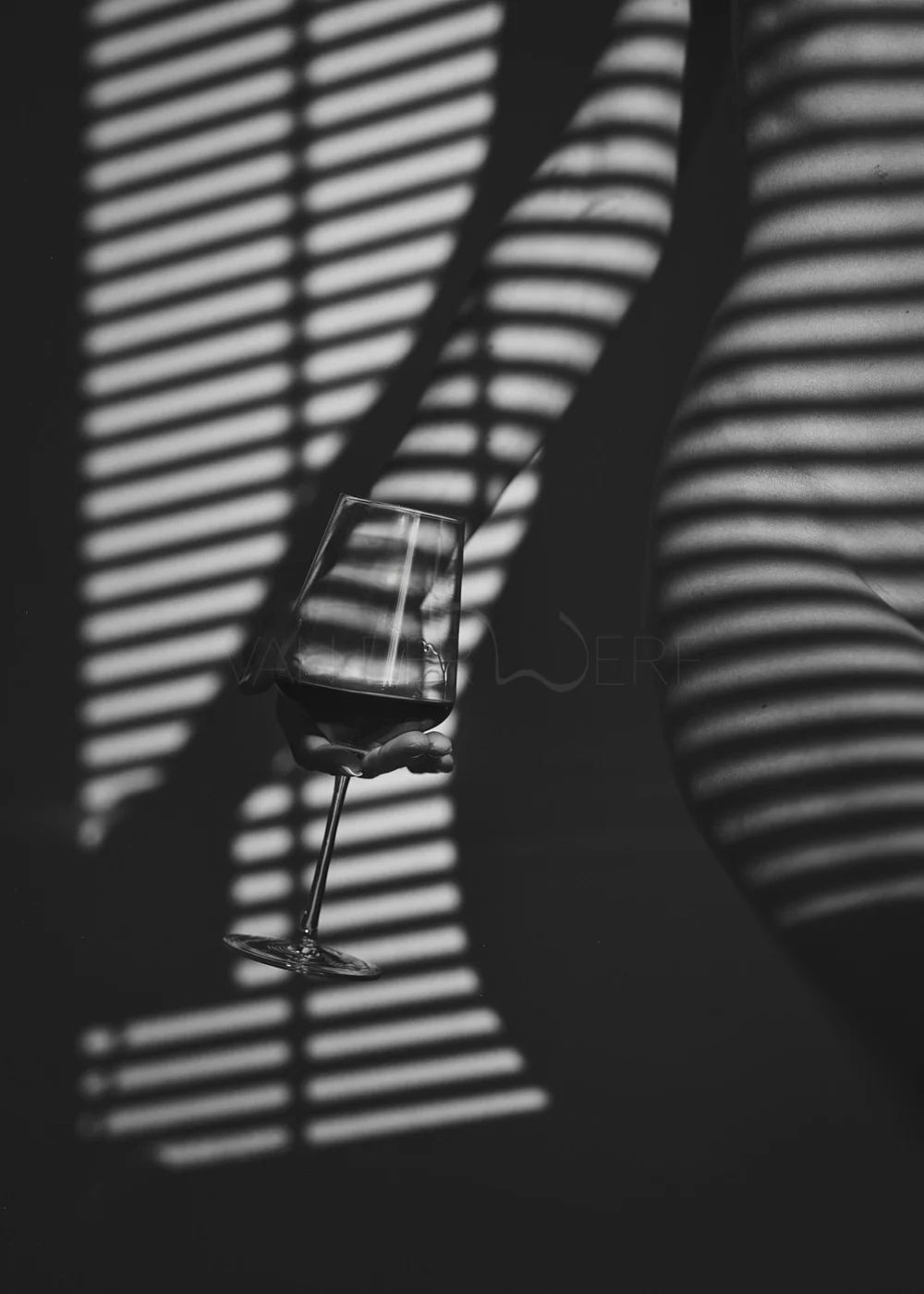 A person holding a wine glass with dark liquid, with striped shadows cast by window blinds creating a pattern on the person's body and the background, in black and white.