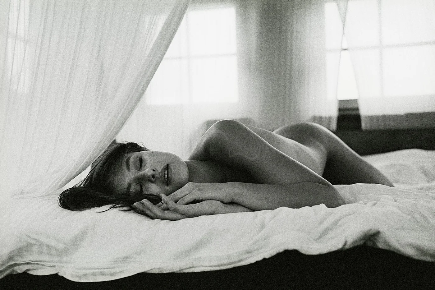 A black and white photo of a woman lying on a bed, partially covered by a sheet, with her eyes closed and a peaceful expression.