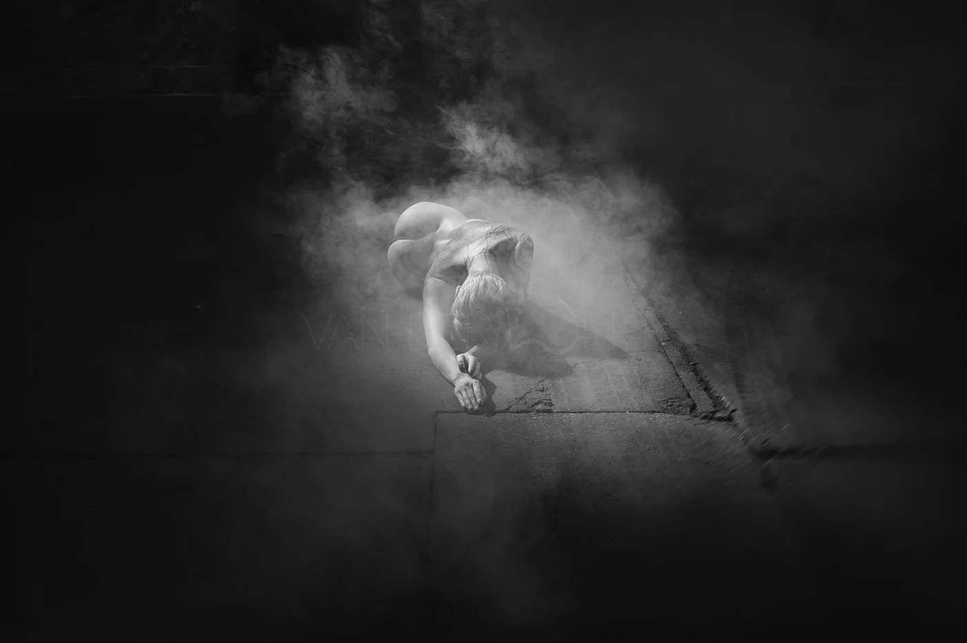 A person crouched down on a street corner with a cloud of smoke or fog around them, in black and white photography.