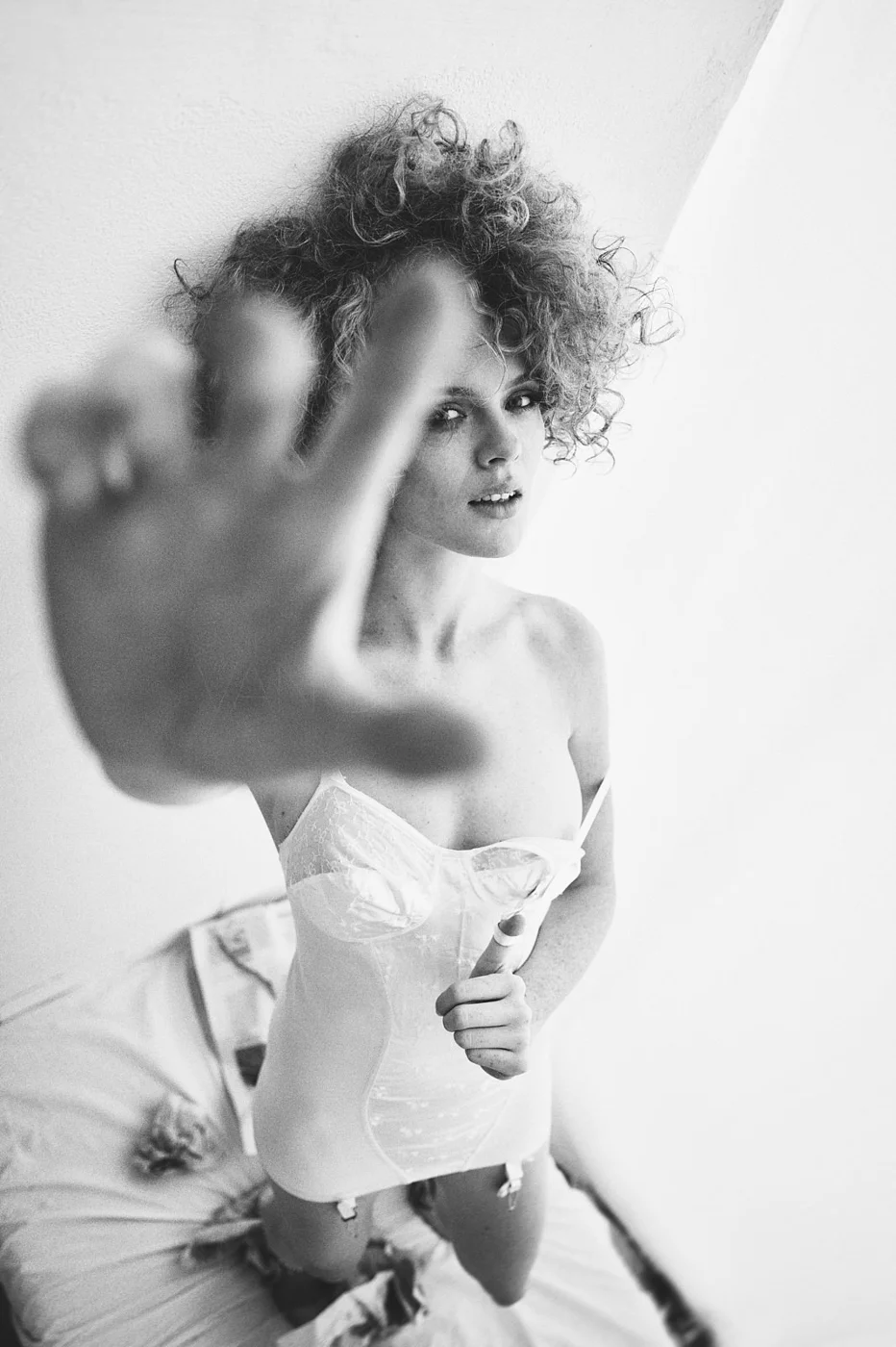 A woman with curly hair reaching towards the camera, wearing a strapless corset and stockings, sitting on a bed in a bright room.