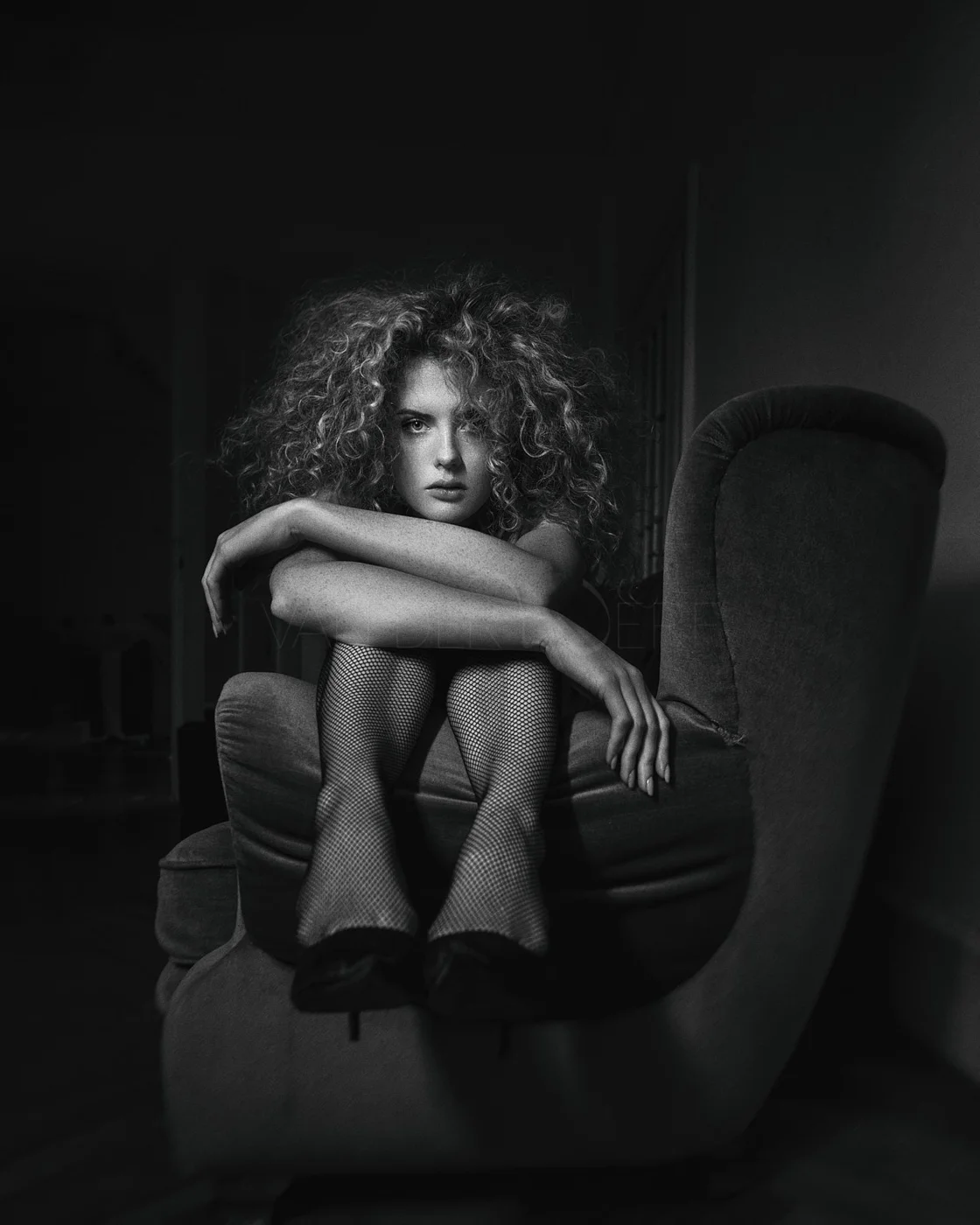 Black and white photo of a young woman with curly hair sitting on an armchair, wearing fishnet stockings and heels, with her arms crossed over her knees, looking directly at the camera.