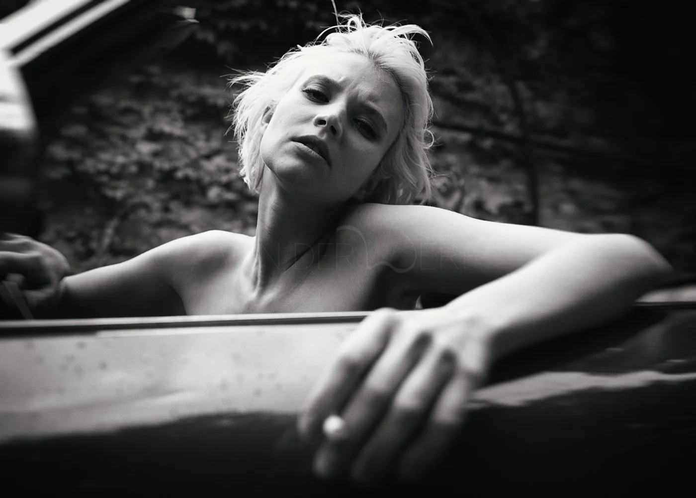 Black and white photo of a woman with short, tousled hair posing topless with her arm resting on a car. She has a serious expression and is looking at the camera, with a blurred outdoor background.
