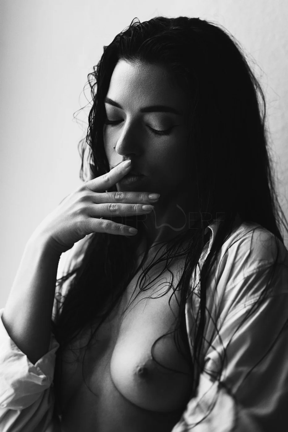 A black-and-white photo of a woman with wet hair, her eyes closed, touching her lips with her hand, partially revealing her shoulder and breast.