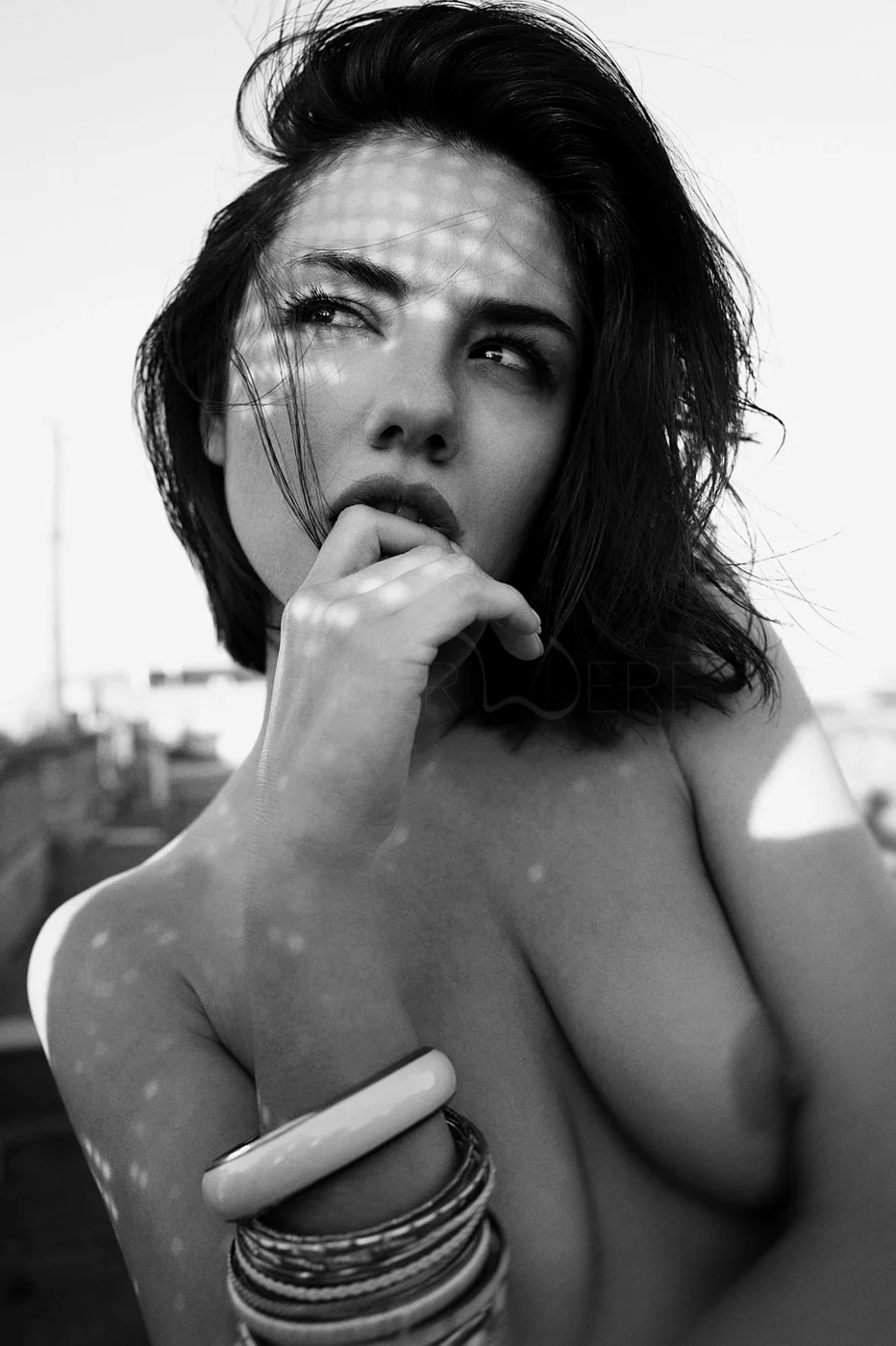 Black and white photograph of a woman with short, dark hair, looking to the side with her finger near her lips, wearing multiple bracelets on her wrist, with sunlight casting a pattern of shadows across her face and shoulder.