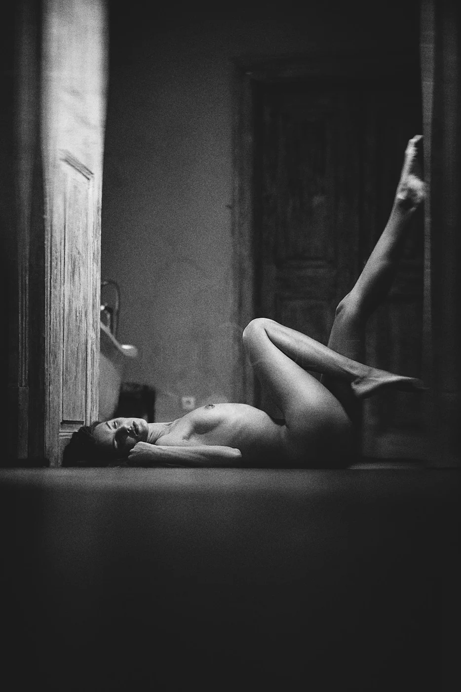 A black and white photo of a nude woman lying on her side on the floor with her head resting on her hand, one leg bent and the other straight, in a dimly lit room with wooden doors.