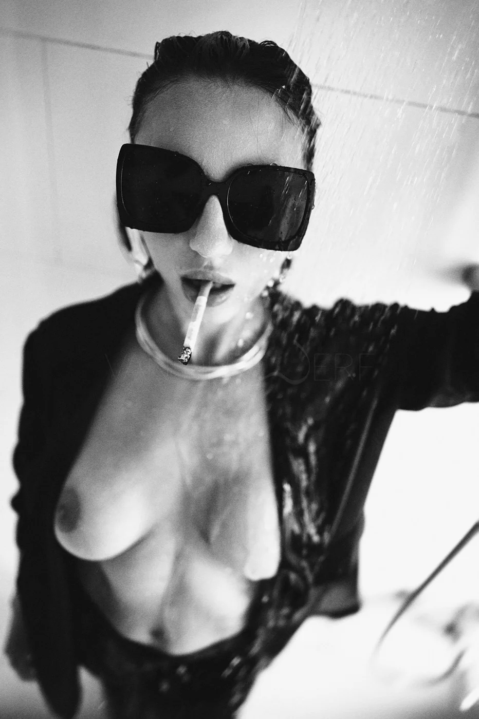 A woman wearing large sunglasses with water and a cigarette in her mouth, partially revealing her chest, dressed in dark clothing.