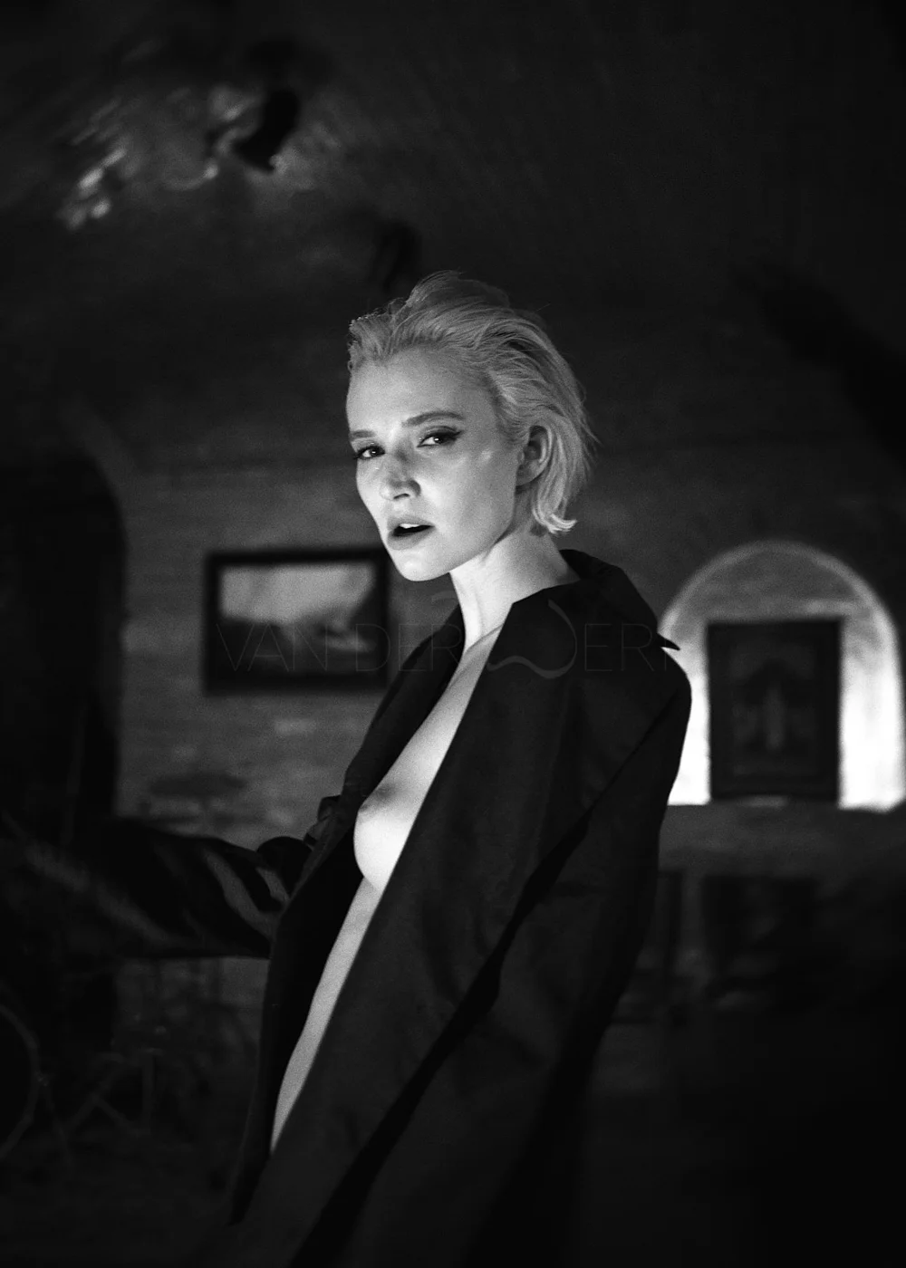 Black and white photograph of a woman with short, styled blonde hair, wearing a partially open blazer exposing her chest, in an indoor setting with framed pictures on the wall.