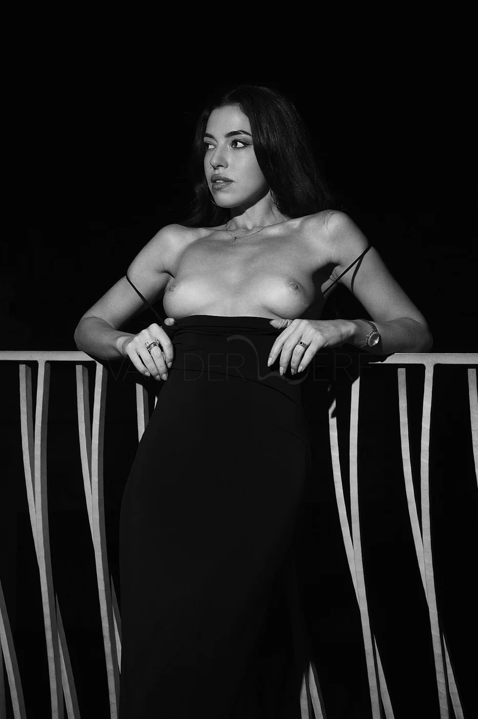 Black and white photo of a woman topless, wearing a long skirt, holding her dress with her hands, standing in front of a decorative railing.